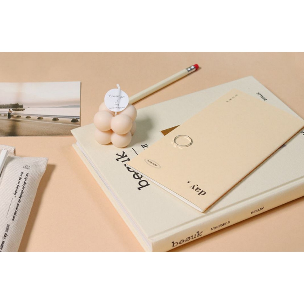 Signature Planner Daily Ivory und Kerze auf einem Buch vor Stiftetui und Foto. Alles in elfenbein, beige farbenen Toenen.