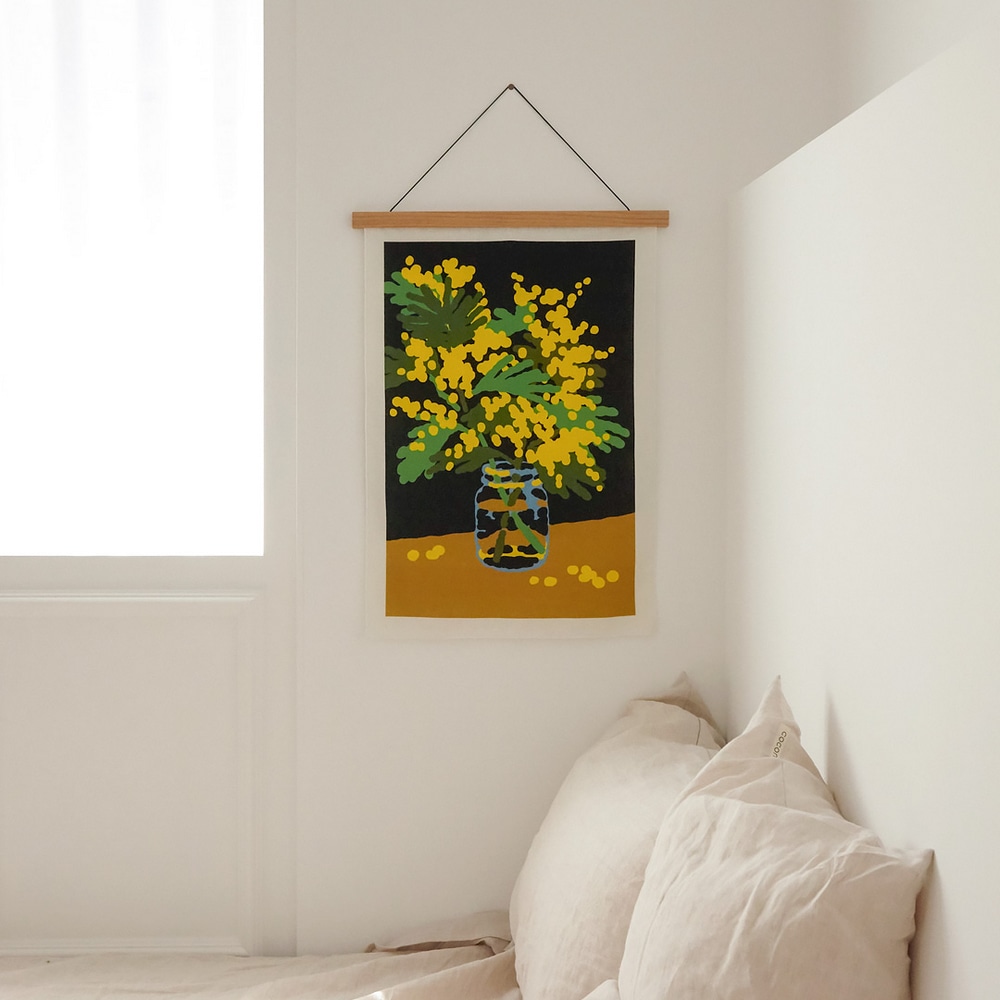 Stoffposter Mimosa, mit gelben Mimosen in einem Einmachglas, am dem Schlafzimmerfenster neben Bettkissen mit beigem Kissenbezug.