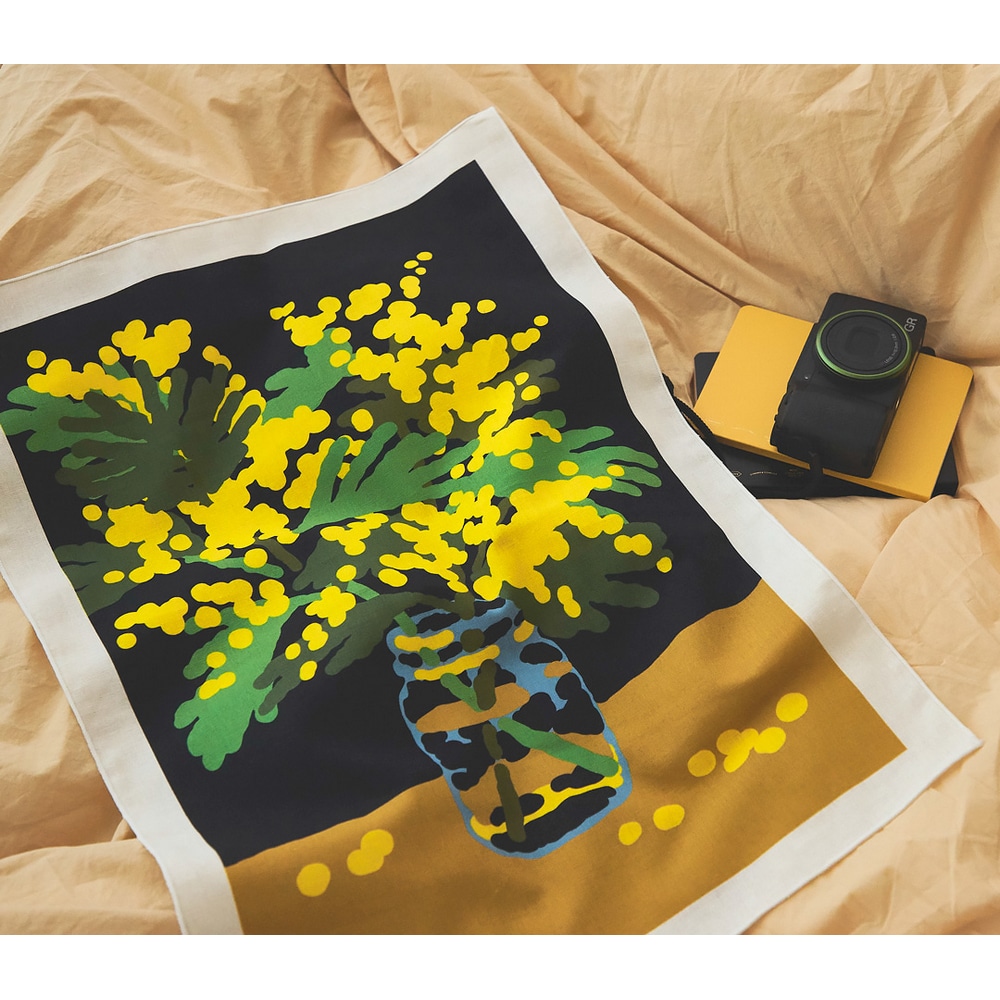 Stoffposter Mimosa, mit gelben Mimosen in einem Einmachglas, auf einem Bett ausgebreitet. Daneben eine schwarze Kompaktkamera.
