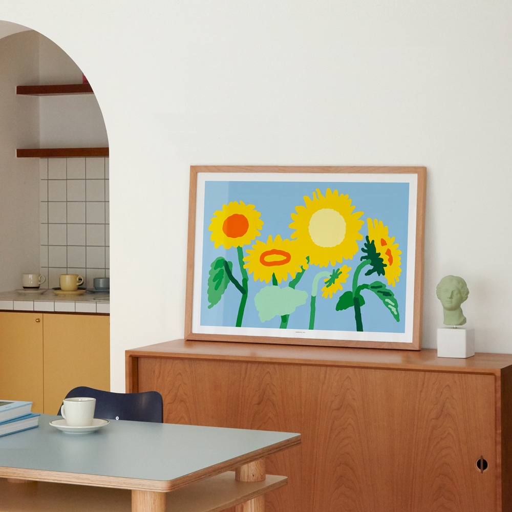 In Holz gerahmter Kunstdruck Sunflower, Sonnenblumen mit gruenen Stengeln und gelben Blueten, auf braunem Sideboard hinter Kuechentisch mit Kaffeetasse.