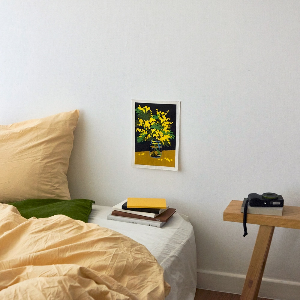 Textilposter Mini Mimosa, mit gelben Mimosen auf schwarzem Hintergrund, an Wand ueber Bett mit gelben Bezuegen gepinnt.